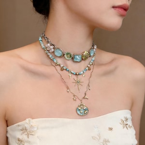Puede incluir: Conjunto de collares de varias capas con varios colgantes y cuentas. La capa superior tiene piedras cuadradas y ovaladas de color azul claro, con detalles florales verdes. La segunda capa tiene cuentas turquesas y pequeñas perlas. La capa inferior tiene un colgante de estrella y concha.