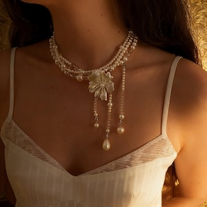 Puede incluir: Un collar de perlas de varias hebras con un gran colgante de flores blancas. El collar presenta detalles colgantes de perlas y cristales. La modelo lleva un vestido blanco de tirantes finos.