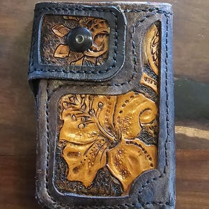 Könnte beinhalten: Handgefertigte Lederbrieftasche mit floralem Design. Die Brieftasche hat eine dunkelbraune und schwarze Farbgebung mit einem Knopfverschluss. Das florale Design ist in einem helleren Braunton gehalten und setzt Akzente.
