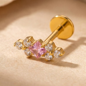 May include: A gold-colored lip piercing stud with a cluster design. The stud features a central square pink gemstone, surrounded by smaller, clear gemstones. The piercing is set against a neutral background.