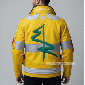 Könnte beinhalten: Gelbe Lederjacke mit grünem Grafikdesign auf dem Rücken. Die Jacke hat silberne Reflexstreifen und das Wort "EMERGENCY" am Kragen. Der Text "HIDE STREET" ist ebenfalls sichtbar.