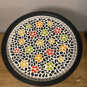 May include: A round mosaic table top with a black border. The mosaic design features colorful flower patterns in red, yellow, orange, and green against a black and white background. The table top is approximately 61 cm in diameter.