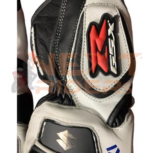 May include: Black, white, and red motorcycle gloves with the Suzuki logo. The gloves feature a leather construction with reinforced padding and stitching. The design includes the letters "GSX" in red.