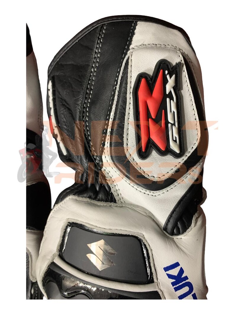 May include: Black, white, and red motorcycle gloves with the Suzuki logo. The gloves feature a leather construction with reinforced padding and stitching. The design includes the letters "GSX" in red.