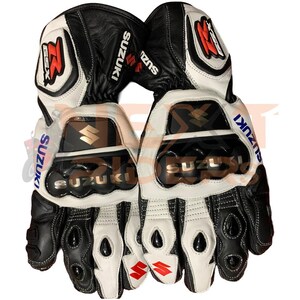 May include: Pair of black and white motorcycle gloves with the Suzuki logo. The gloves have a leather exterior with protective padding on the knuckles and palms. The Suzuki logo is displayed in multiple locations on the gloves.