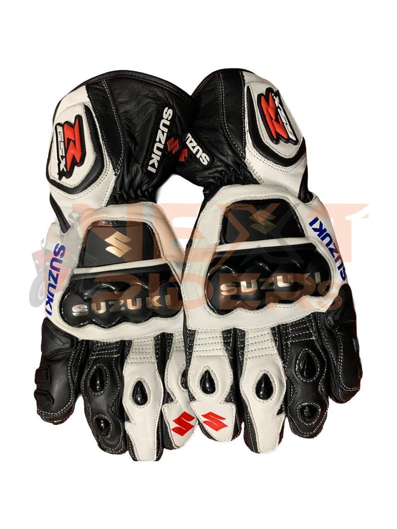 May include: Pair of black and white motorcycle gloves with the Suzuki logo. The gloves have a leather exterior with protective padding on the knuckles and palms. The Suzuki logo is displayed in multiple locations on the gloves.