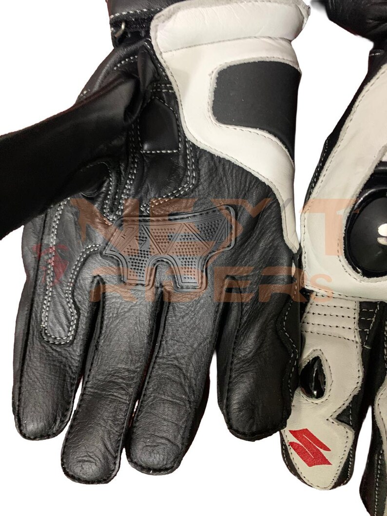 May include: Black and white motorcycle gloves with reinforced knuckles and palm padding. The gloves feature a leather construction with white accents and stitching. The gloves have a velcro strap closure and a Suzuki logo.