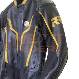 Op de afbeelding: Zwarte leren motorjas met gele accenten en de letter "R" op de mouw. De jas heeft een ronde patch met een wit en geel ontwerp. De jas hangt aan een hanger.