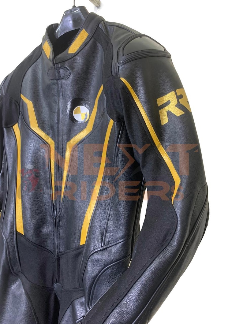Op de afbeelding: Zwarte leren motorjas met gele accenten en de letter "R" op de mouw. De jas heeft een ronde patch met een wit en geel ontwerp. De jas hangt aan een hanger.