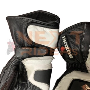 May include: Black and white motorcycle gloves with the Suzuki logo. The gloves feature white accents on the back of the hand and wrist, with a black leather exterior and white stitching. The gloves have a strap closure.
