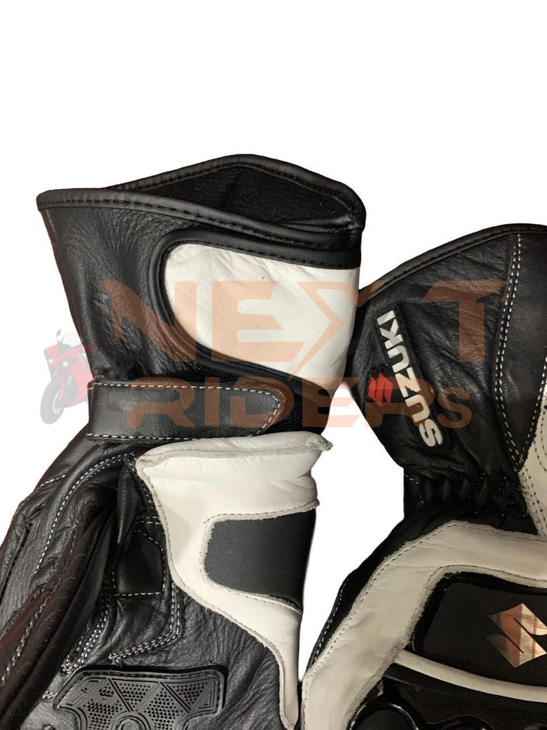 May include: Black and white motorcycle gloves with the Suzuki logo. The gloves feature white accents on the back of the hand and wrist, with a black leather exterior and white stitching. The gloves have a strap closure.