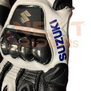 May include: Close-up of a white and black motorcycle glove with the word "SUZUKI" in blue. The glove has a protective knuckle guard and the Suzuki logo.