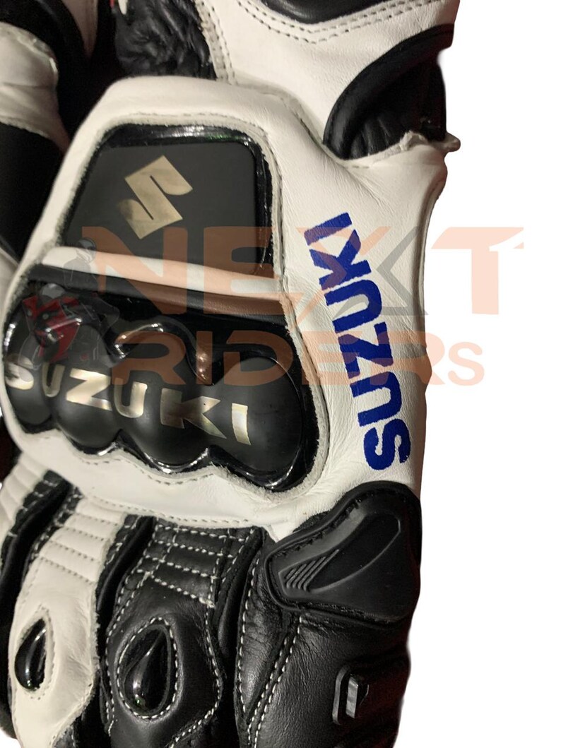 May include: Close-up of a white and black motorcycle glove with the word "SUZUKI" in blue. The glove has a protective knuckle guard and the Suzuki logo.