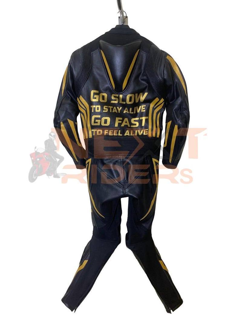 Op de afbeelding: Zwart en goud motorracepak. Het pak heeft gouden accenten en de tekst "Go Slow To Stay Alive, Go Fast To Feel Alive" op de rug. Het pak is gemaakt van leer en heeft een tweedelig ontwerp.
