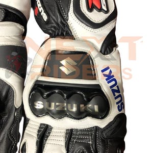 May include: Black, white, and gray motorcycle gloves with the Suzuki logo. The gloves have reinforced knuckles and the word "SUZUKI" in silver lettering. The gloves are made of leather and have a sporty design.