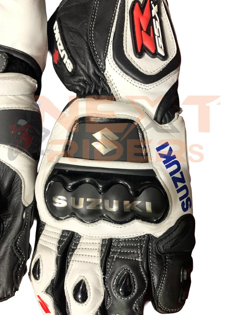 May include: Black, white, and gray motorcycle gloves with the Suzuki logo. The gloves have reinforced knuckles and the word "SUZUKI" in silver lettering. The gloves are made of leather and have a sporty design.