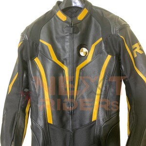 Op de afbeelding: Zwart leren motorpak met gele accenten. Het pak heeft een lange ritssluiting, gewatteerde schouders en een rond embleem op de borst. Het pak hangt aan een hanger.