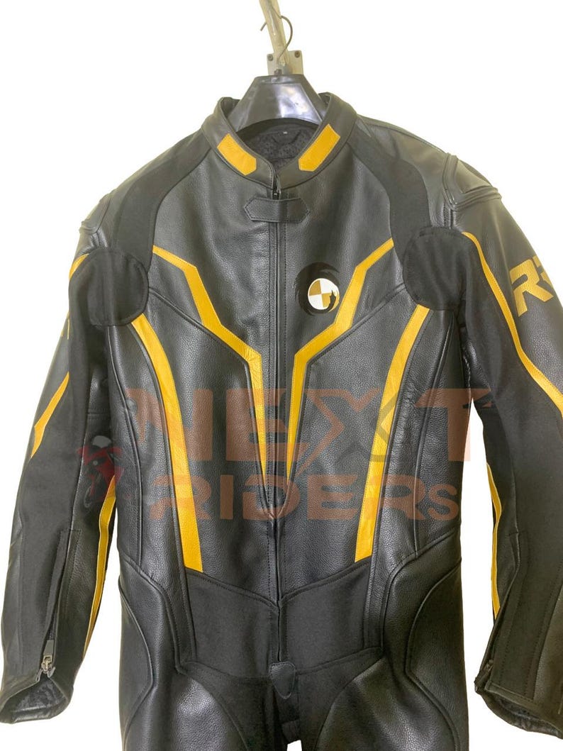 Op de afbeelding: Zwart leren motorpak met gele accenten. Het pak heeft een lange ritssluiting, gewatteerde schouders en een rond embleem op de borst. Het pak hangt aan een hanger.