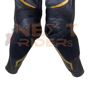 Op de afbeelding: Zwarte leren motorbroek met gouden accenten. De broek heeft versterkte knie- en scheenbeenstukken, met de tekst "NEXT RIDERS" zichtbaar. Het ontwerp omvat gouden strepen en panelen, en zwarte klittenbandsluitingen.