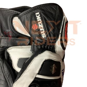 May include: Black and white motorcycle gloves with the Suzuki logo. The gloves have a leather exterior with white accents and stitching. The Suzuki logo is red and black.