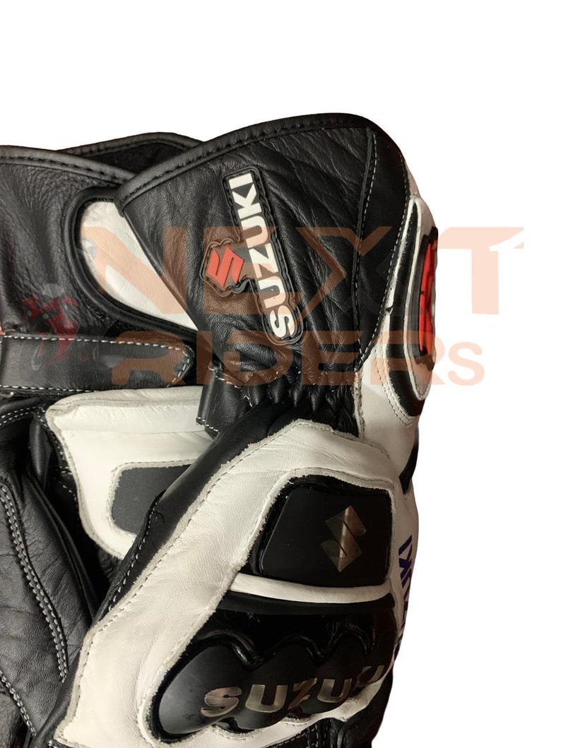 May include: Black and white motorcycle gloves with the Suzuki logo. The gloves have a leather exterior with white accents and stitching. The Suzuki logo is red and black.