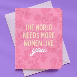 May include: A pink greeting card with a floral pattern and the text "THE WORLD NEEDS MORE WOMEN LIKE YOU" in white and yellow. The card is inside a white envelope, set against a purple background.