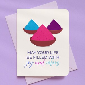 May include: Greeting card with three bowls of colorful powder in blue, pink, and purple. The card reads "MAY YOUR LIFE BE FILLED WITH joy and colors" in a gradient font. The card is on a purple background.