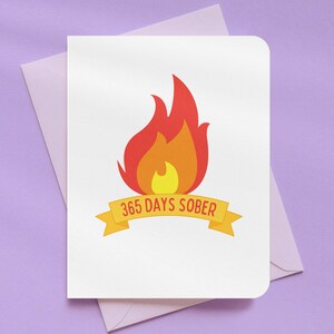 May include: A white greeting card with a stylized flame graphic in red, orange, and yellow. A yellow banner below the flame reads "365 DAYS SOBER." The card is inside a lavender envelope, set against a purple background.