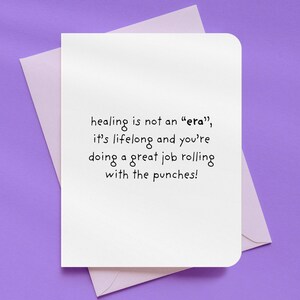 May include: A white greeting card with rounded corners, featuring the text "healing is not an 'era', it's lifelong and you're doing a great job rolling with the punches!" The card is inside a light purple envelope, set against a purple background.