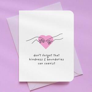 May include: A white greeting card with a pink heart and two hands reaching towards each other. The card has the text "don't forget that kindness & boundaries can coexist". The card is inside a white envelope, all on a purple background.