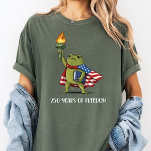 Puede incluir: Una camiseta verde oliva con el gráfico de una rana verde vestida como la Estatua de la Libertad, sosteniendo una antorcha encendida y un libro azul, con una capa con los colores de la bandera estadounidense. El texto 250 YEARS OF FREEDOM aparece debajo.