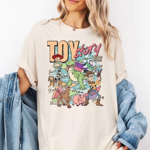 May include: A cream-colored short-sleeve t-shirt featuring a graphic print of Toy Story characters including Woody, Buzz Lightyear, Jessie, Rex, Slinky Dog, Bullseye, and Hamm with the text Toy Story Since 1995 and individual character names.