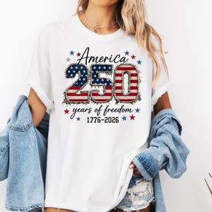 May include: A white short-sleeved t-shirt featuring a graphic with the text America 250 years of freedom 1776-2026. The numbers 250 are filled with a red, white, and blue American flag pattern and surrounded by small red and blue stars.