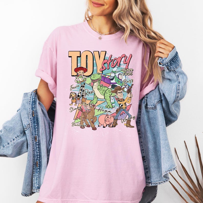 May include: A light pink t-shirt featuring a graphic print of Toy Story characters including Woody, Buzz Lightyear, Jessie, Rex, Slinky Dog, Bullseye, and Hamm. The text reads TOY story Since 1995, with character names written in cursive script.
