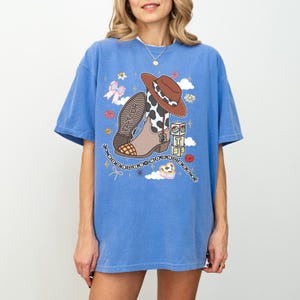 May include: A blue short-sleeve t-shirt featuring a graphic print of a brown cowboy hat, a cow-print western boot with the number 13, a stack of blocks with the letters T, S, and a beaded chain with the text Taylor Swift in black lettering.
