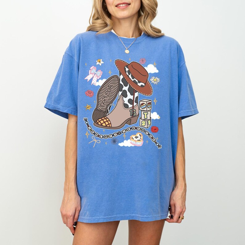 May include: A blue short-sleeve t-shirt featuring a graphic print of a brown cowboy hat, a cow-print western boot with the number 13, a stack of blocks with the letters T, S, and a beaded chain with the text Taylor Swift in black lettering.