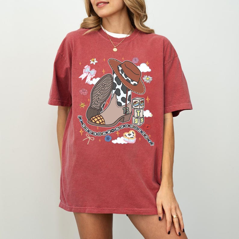 May include: A brick red short-sleeve t-shirt featuring a graphic print of a brown and white cow print cowboy boot, a brown cowboy hat, three stacked blocks with the letters T, S, and a heart, a bow, and a beaded string with the text Taylor.