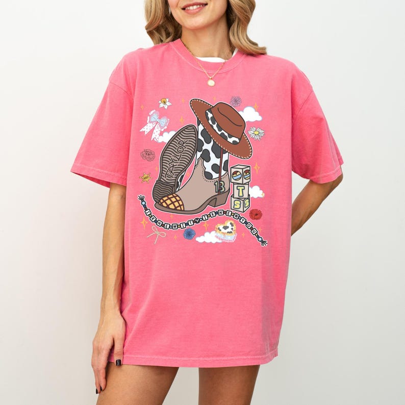 May include: A bright pink short-sleeve t-shirt featuring a graphic print of a brown cowboy hat, a cow-print western boot with the number 13, and a stack of blocks with the letters T, S, and T. The design includes flowers, stars, and a beaded chain.