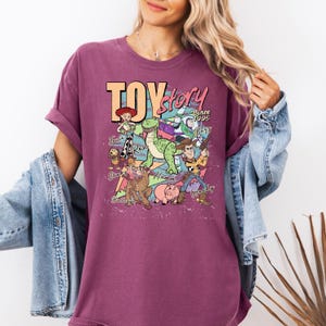 May include: A maroon short-sleeve t-shirt featuring a graphic print of Toy Story characters including Woody, Buzz Lightyear, Jessie, Rex, Slinky Dog, Bullseye, and Hamm with the text Toy Story Since 1995 and individual character names printed in a retro style.