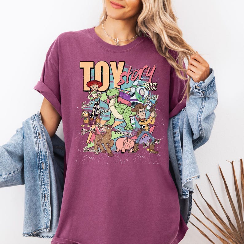May include: A maroon short-sleeve t-shirt featuring a graphic print of Toy Story characters including Woody, Buzz Lightyear, Jessie, Rex, Slinky Dog, Bullseye, and Hamm with the text Toy Story Since 1995 and individual character names printed in a retro style.
