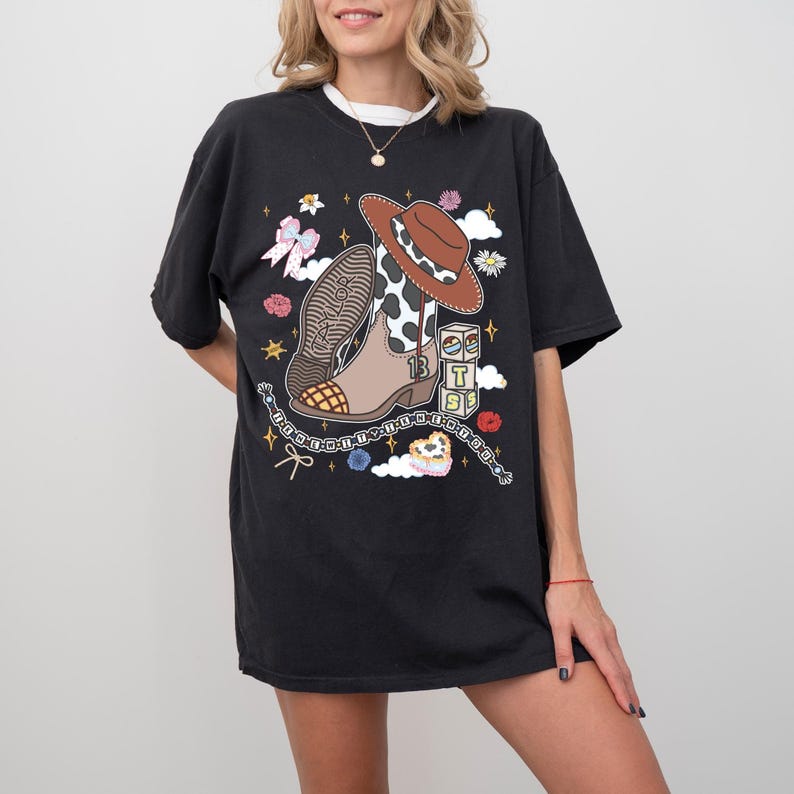May include: A black short-sleeve t-shirt featuring a graphic print of a brown cowboy boot with a cow-print shaft, a brown cowboy hat, a bow, small flowers, stars, a heart, and stacked blocks with the letters T, S, and the number 13.