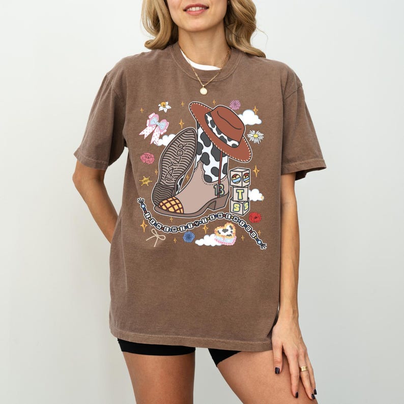 May include: A brown short-sleeve t-shirt featuring a graphic print of a cowboy boot with a cow print pattern, a brown cowboy hat, a stack of blocks with the letters T, S, and S, a bow, flowers, stars, and a beaded chain with the text Taylor.