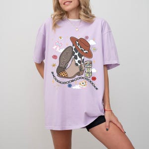 May include: A light purple short-sleeve t-shirt featuring a graphic print of a brown cowboy hat, a cow-print boot with the number 13, stacked blocks spelling TS, a beaded necklace, a pink bow, flowers, and clouds on the front center.