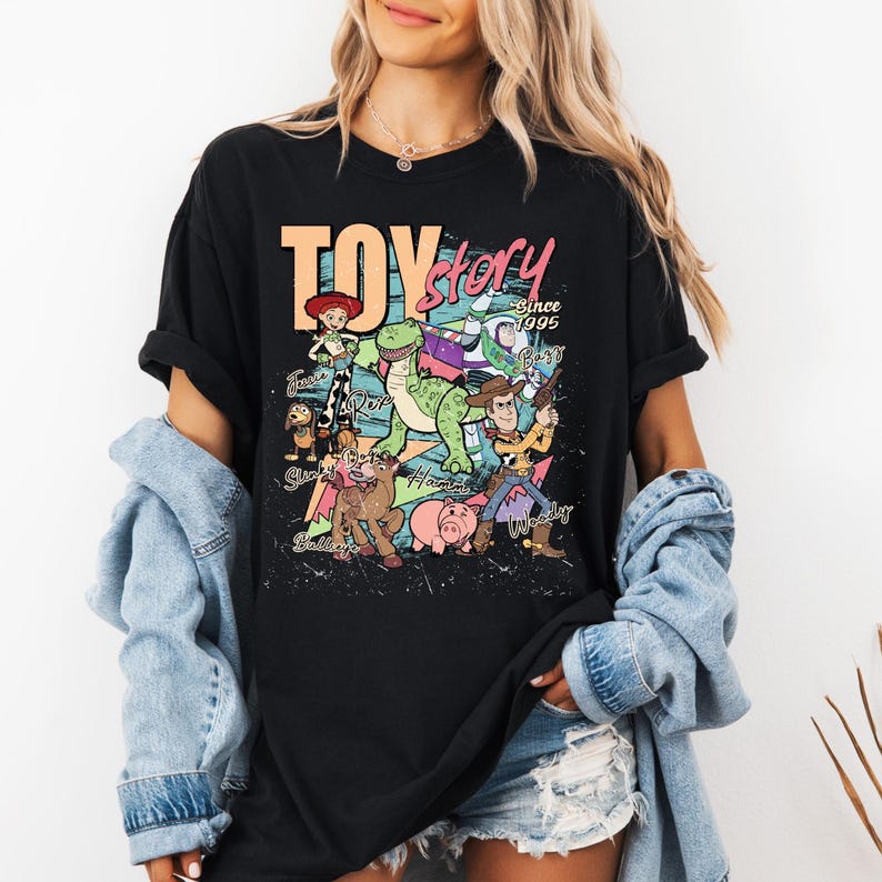 May include: A black short-sleeve t-shirt featuring a colorful graphic print of Toy Story characters including Woody, Buzz Lightyear, Rex, and Jessie. The text reads TOY story Since 1995 with character names written in cursive script below each figure.