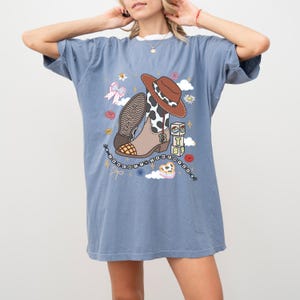 May include: A blue short-sleeve t-shirt featuring a graphic print of a brown cowboy hat, a cow-print boot with the number 13, alphabet blocks spelling TS, a beaded necklace, a pink bow, flowers, and clouds with the text Taylor Swift.