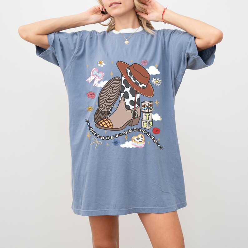 May include: A blue short-sleeve t-shirt featuring a graphic print of a brown cowboy hat, a cow-print boot with the number 13, alphabet blocks spelling TS, a beaded necklace, a pink bow, flowers, and clouds with the text Taylor Swift.