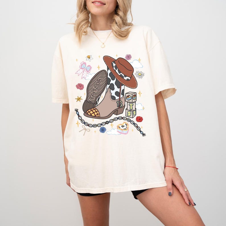 May include: An off-white short-sleeve t-shirt featuring a graphic print of a brown cowboy boot with a cow-print shaft, a brown cowboy hat, a beaded bracelet, and three stacked blocks labeled with the letters T, S, and the number 13.