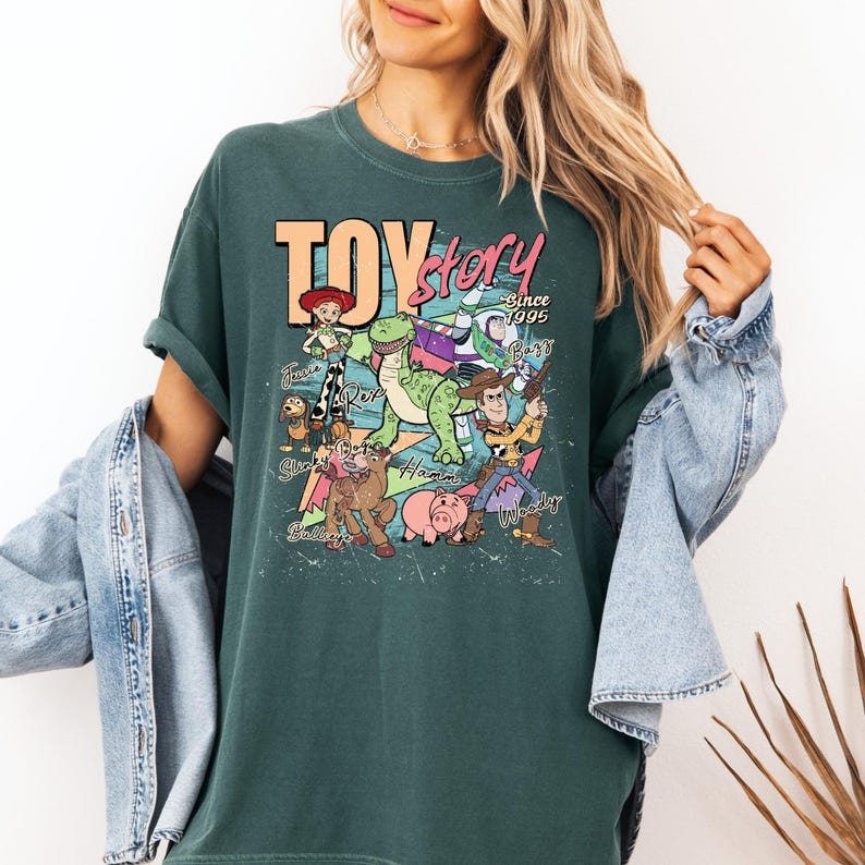 May include: A dark green short-sleeve t-shirt featuring a graphic print of Toy Story characters including Woody, Buzz Lightyear, Jessie, Rex, Slinky Dog, and Hamm. The text reads Toy Story Since 1995 with character names written in cursive script.