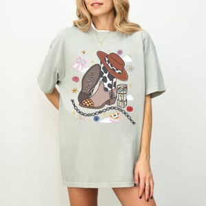May include: A light sage green t-shirt featuring a graphic print of a brown cowboy boot with a cow print shaft, a brown cowboy hat, a beaded bracelet, and stacked blocks with the letters T, S, and the number 13. The boot has the name Taylor.