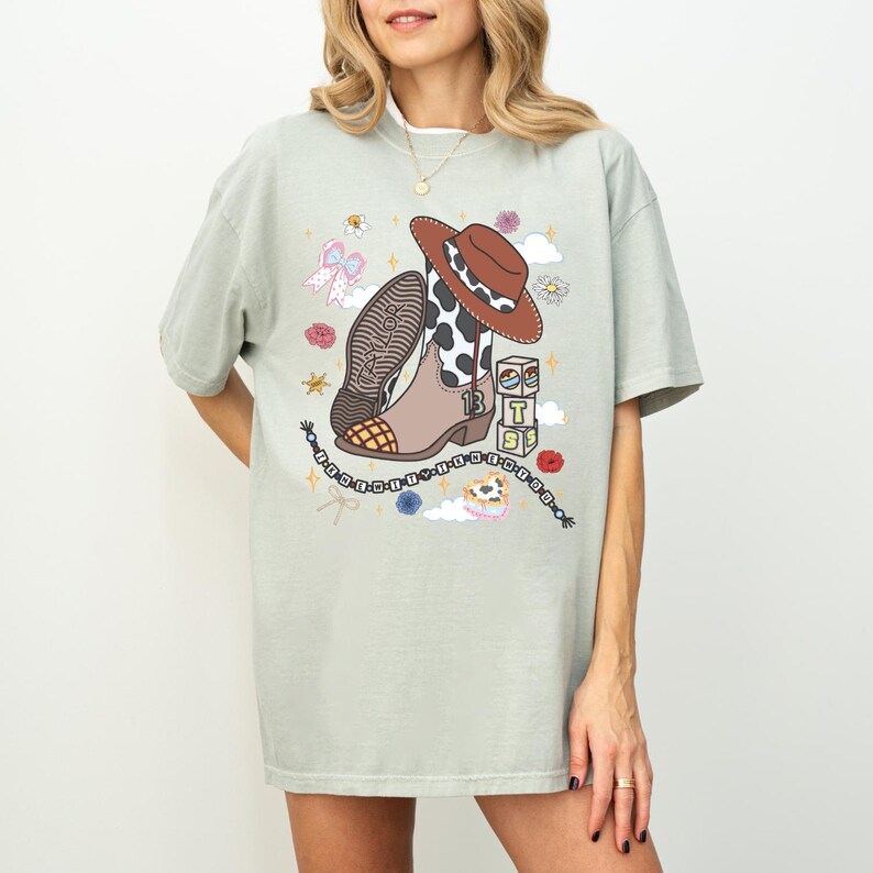 May include: A light sage green t-shirt featuring a graphic print of a brown cowboy boot with a cow print shaft, a brown cowboy hat, a beaded bracelet, and stacked blocks with the letters T, S, and the number 13. The boot has the name Taylor.